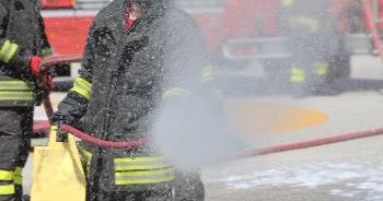 Firefighter in full gear using a fire hose to spray water while on the scene, a fire truck behind them